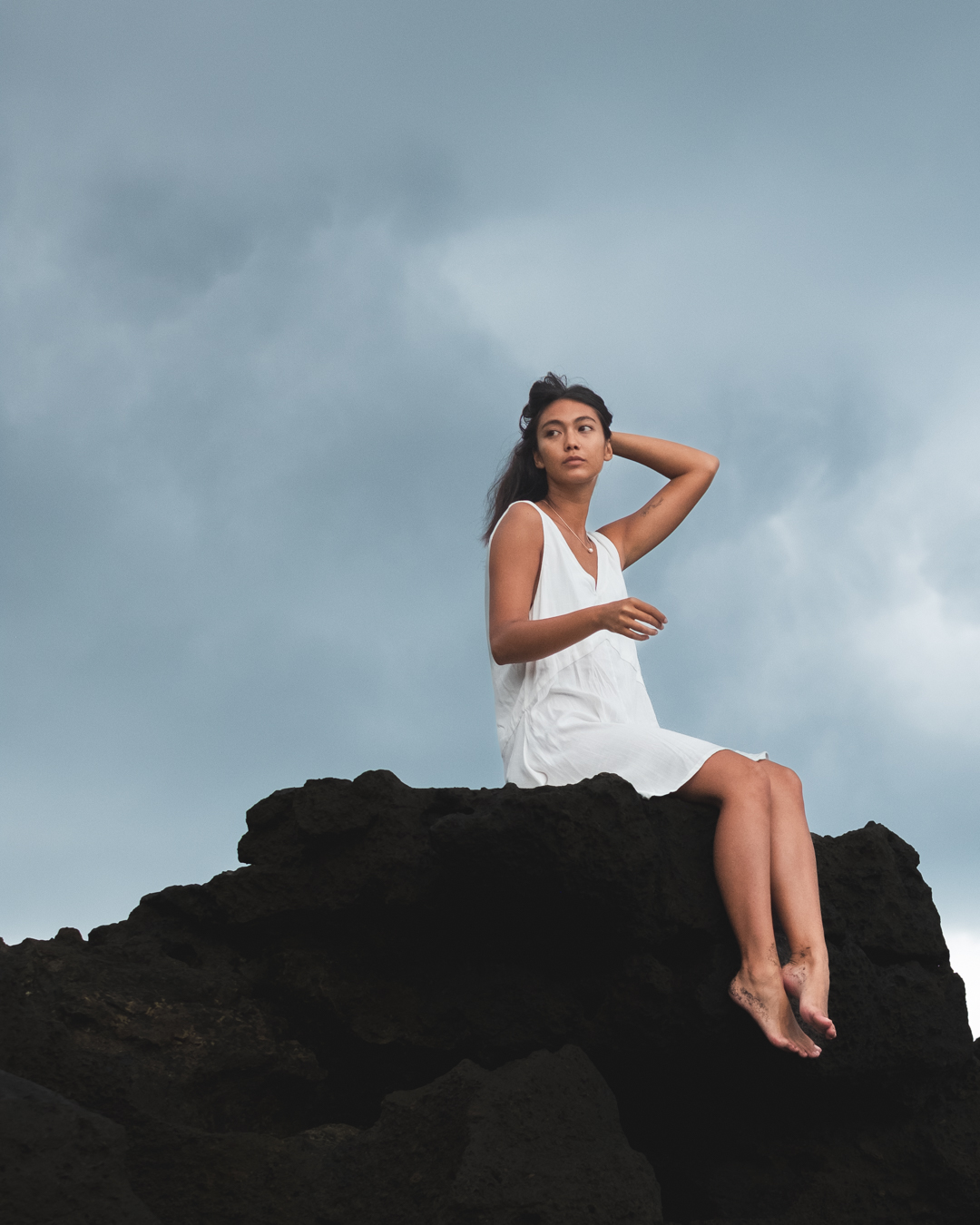 Portrait of a female model in a white sleeveless dress sitting on dark rocks under a cloudy sky, looking off into the distance with one hand resting behind her head.